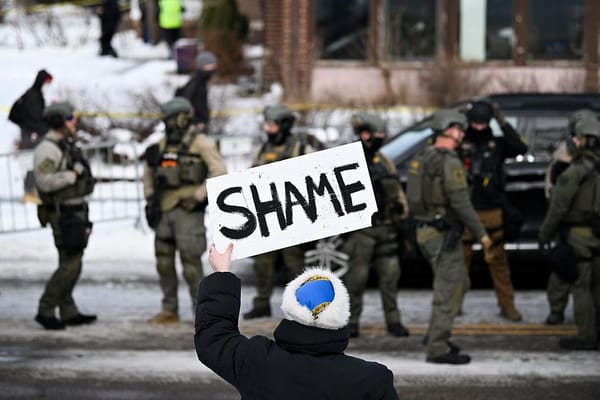 An onlooker holds a sign that reads "Shame" as members of law enforcement work the scene following a suspected shooting by an ICE agent during federal law enforcement operations.